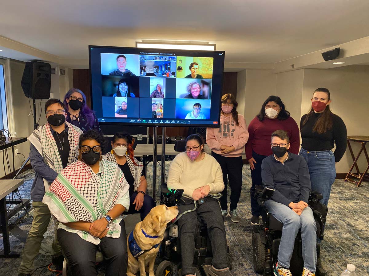 US Alliance members, some using wheelchairs and some standing, pose together in front of a large screen showing others joining virtually during the in-person convening in Washington, DC.