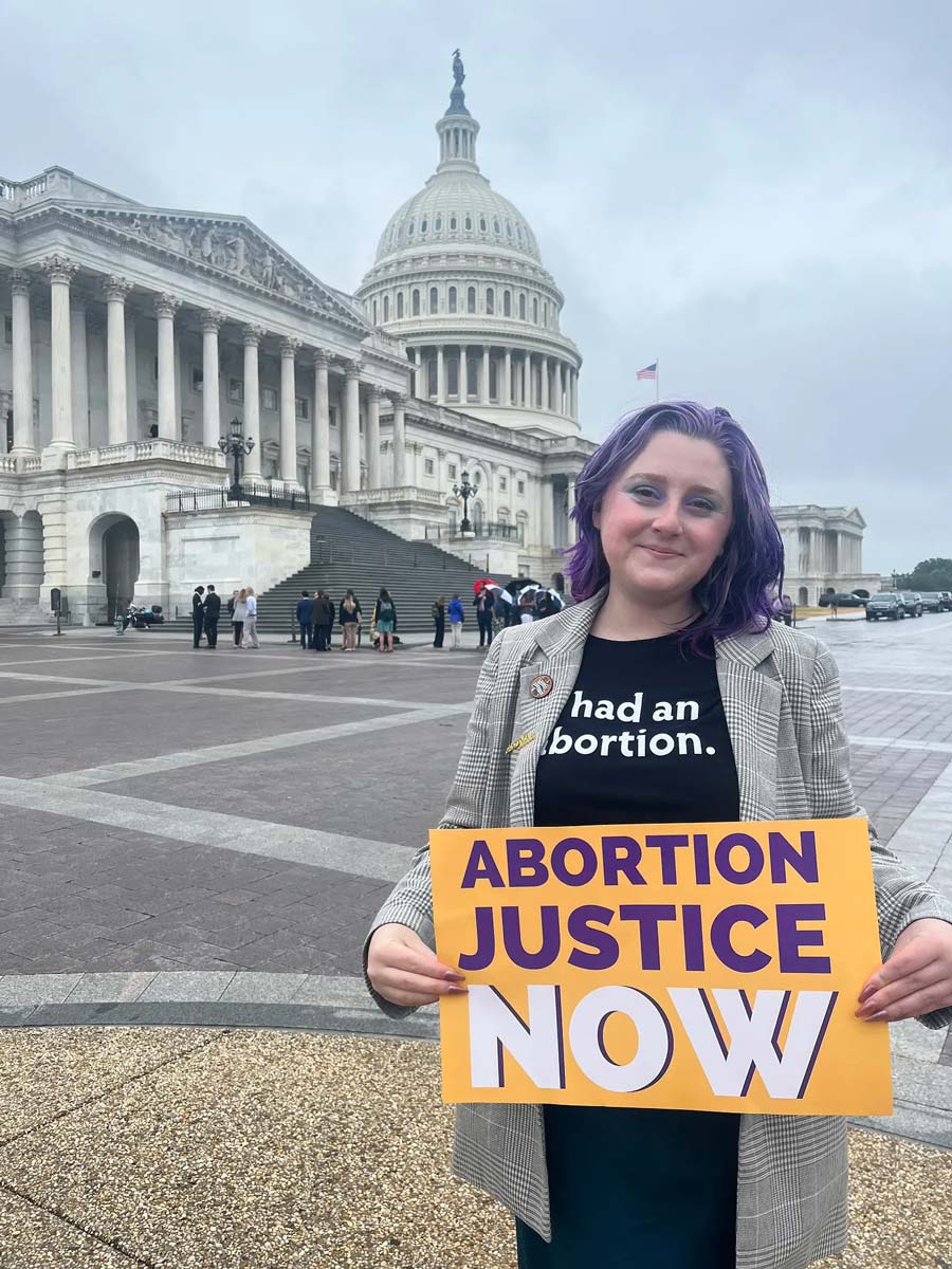 A person with purple hair, wearing a T-shirt that reads “ had an abortion,” and holding a sign that says “ABORTION JUSTICE NOW. The Capital-building is in the background.