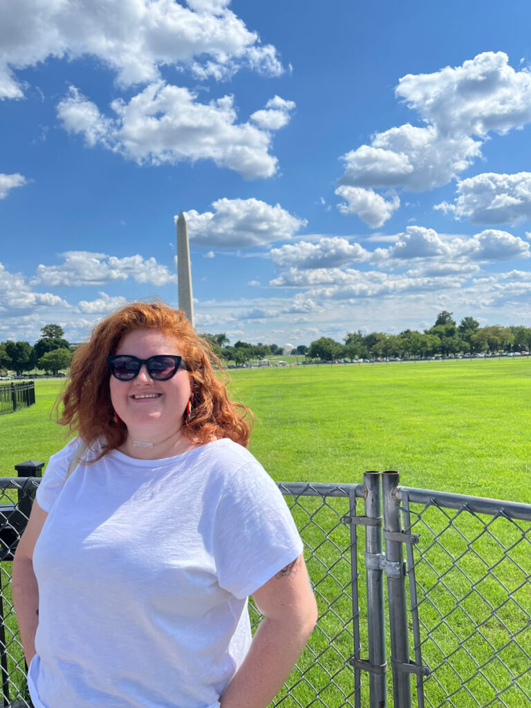 Annabelle, a white woman with medium length red hair, is outside on a sunny day. She is posed in front of a fence at the Ellipse in Washington D.C. with the Washington Monument in the background. She is smiling and is wearing a white t-shirt and dark sunglasses.