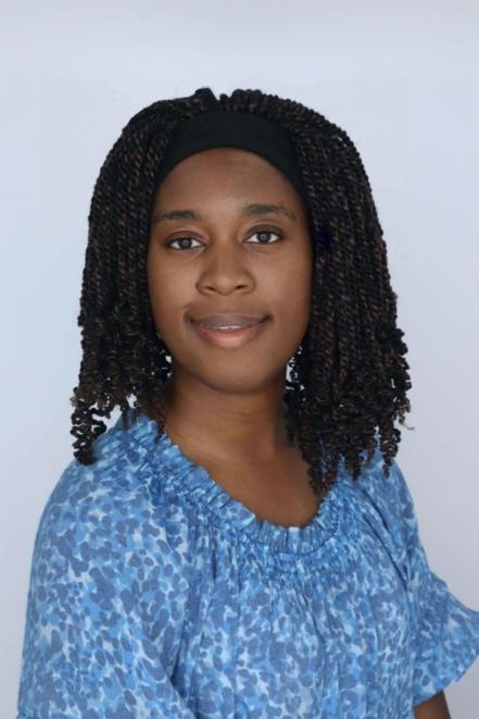 An African American person with brown skin and dark, medium-length coiled twists held back by a black headband. They are wearing a blue blouse with a textured leopard-like pattern in various shades of blue. They look directly at the camera with a soft smile, and the plain white background keeps the focus on their face and expression