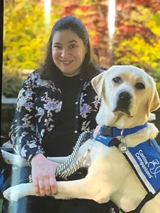 Photo of Janie (she/ her). She is a White and Hispanic woman with shoulder length brown hair. She is wearing a black button- up shirt with multicolored flowers printed on it. She is seated in her power wheelchair and her service dog, Neville has his two front paws across her lap. Neville is a yellow lab golden retriever cross, and is wearing a blue and white service dog vest. The vest inscribed with the words “Canine Companions.” There are trees and greenery in the background of the photo.