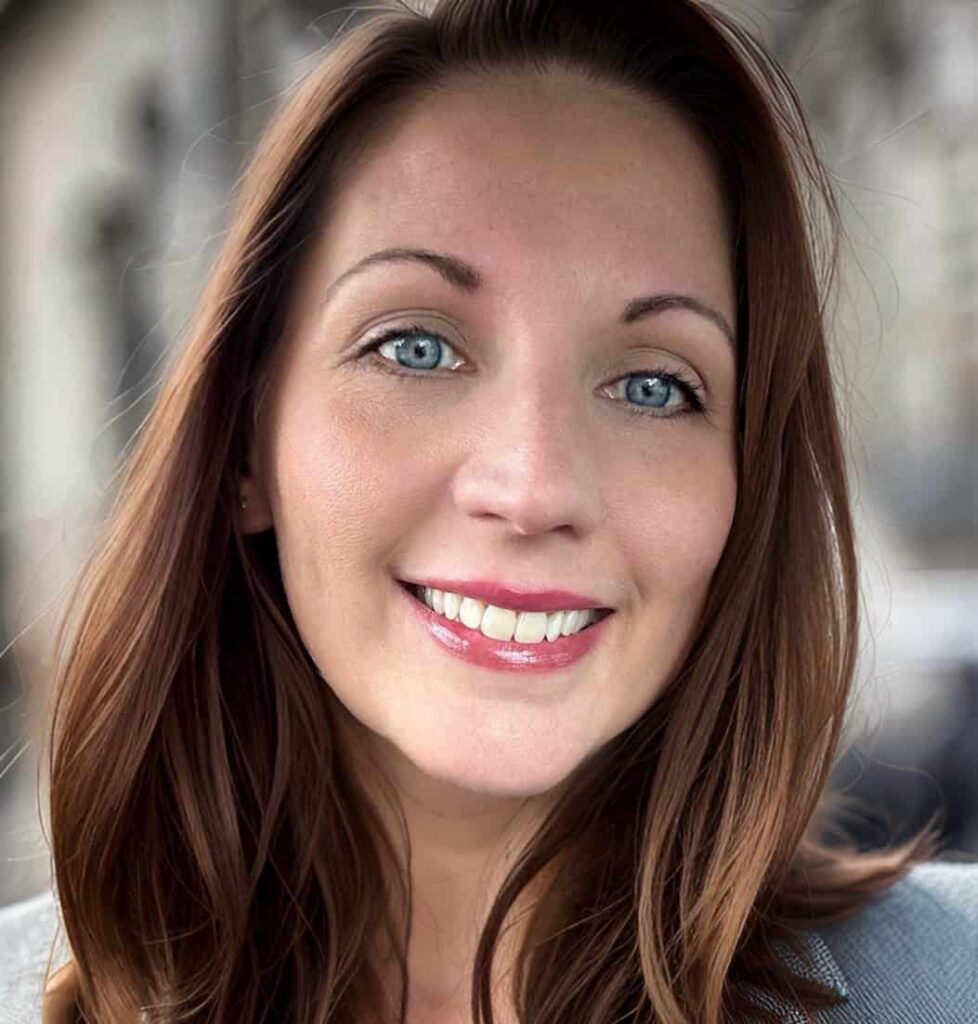 Jessica is a white woman with blue eyes and brown hair and she is smiling at the camera in front of a blurred background. She is wearing a gray suit jacket.