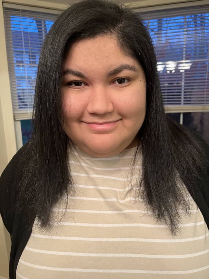 Photo of Kayla Rodriguez (She/they), light skinned Puerto Rican woman and non-binary person with straight black hair going down to the upper half of her chest, black eyebrows, black-like dark brown eyes, and pink lips. Wearing a black sweater on top of a tan shirt that has white stripes on it. The background of this photo has windows behind her.