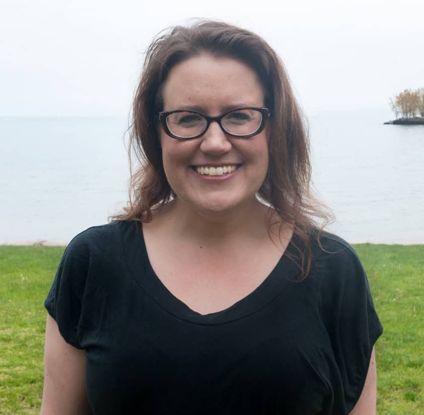 A white cis-gender woman with dark-rimmed glasses and shoulder-length light brown hair smiles at the camera. She wears a black tee shirt with a view of Lake Michigan in the background.