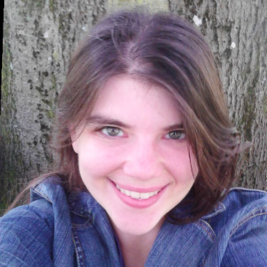 Alicia, a white woman with medium-length dark brown hair and hazel eyes, is smiling at the camera. She is wearing a denim jacket and is sitting in front of a tree where only the bark of the tree is visible.