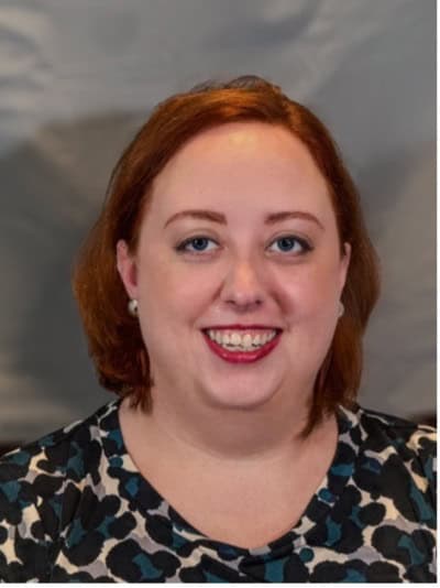 Picture of a white woman with red short red hair cut into a bob. She is sitting in front of a grey background. She is wearing a navy, teal, cream, and black leopard print shirt. She has on large, round, silver earrings and is looking at the camera smiling.