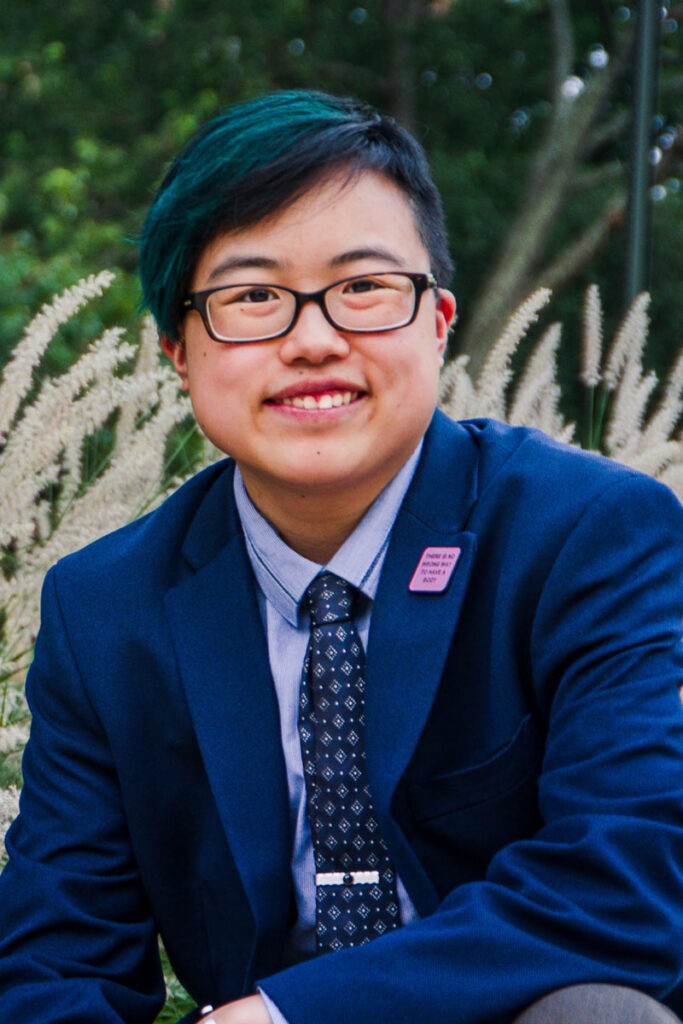 Photo by Sarah Tundermann: Lydia X. Z. Brown smiles in front of reeds blowing in a gentle breeze. They are an East Asian nonbinary/transmasc person with short black hair and glasses, wearing a dark blue suit and blue paisley necktie.