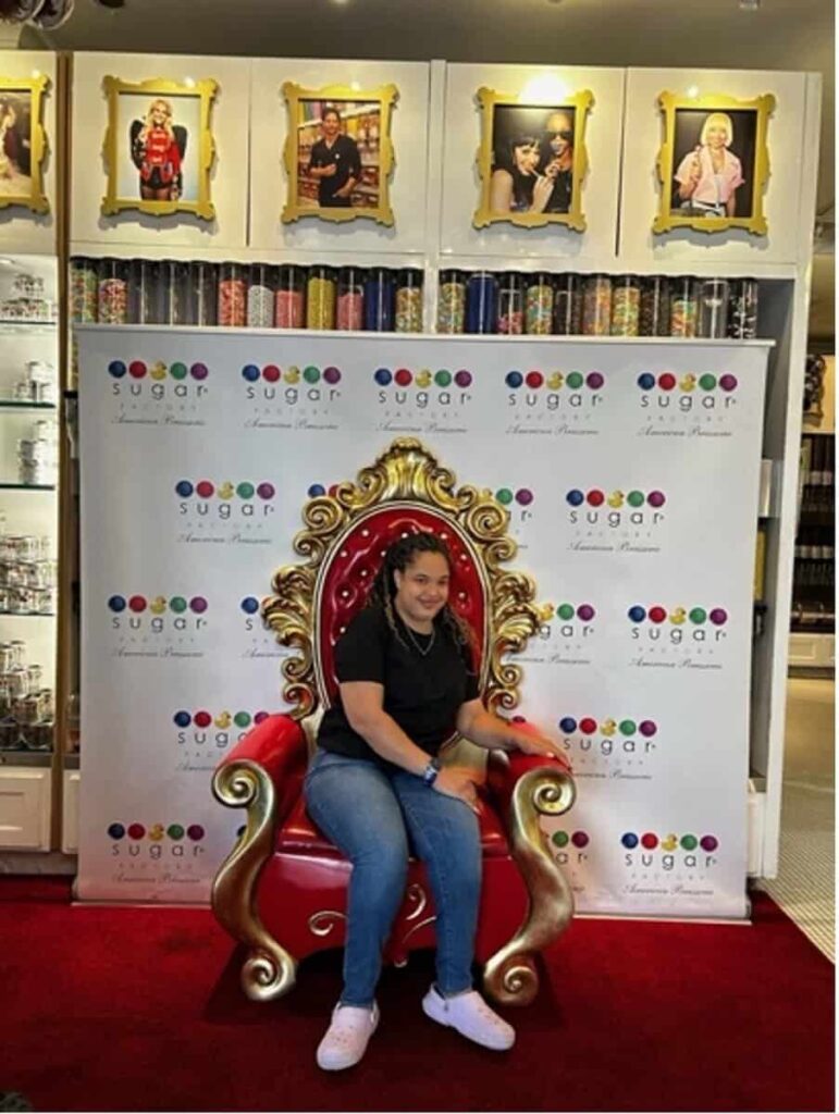 The image is of Saphire during her travels last year to Chicago in the Sugar Factory. In the image, Saphire is a light bi-racial woman wearing medium-washed jeans, a black tee, and lavender croc shoes. Saphire is sitting in a red royal-style chair with gold accents and a red carpet with a background behind which is white with multiple-colored candies and the Sugar Factory written on it in black. Above Saphire, in the background, are various colored candies and golden frames with different celebrities on the wall.